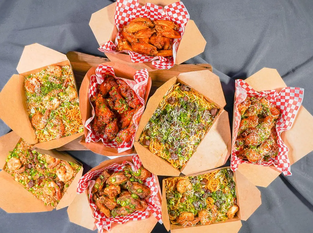 Overhead view of Nghbrs Kitchen takeout boxes on red-checkered paper: saucy wings, garlic noodles with shrimp, and fried rice with shrimp and green onion