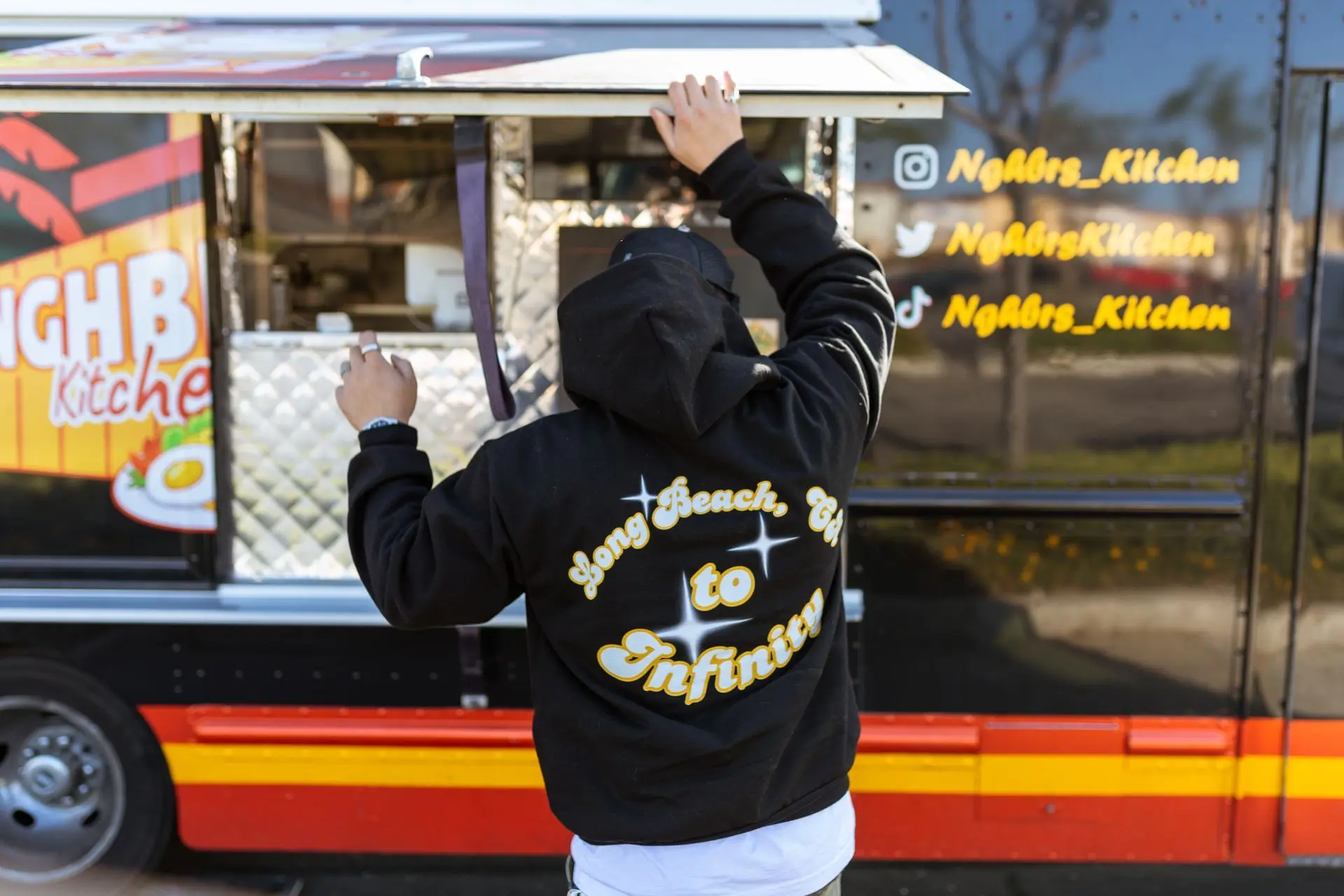A person in a black hoodie opening the service window of the black NGHBRS Kitchen food truck with red and gold stripes