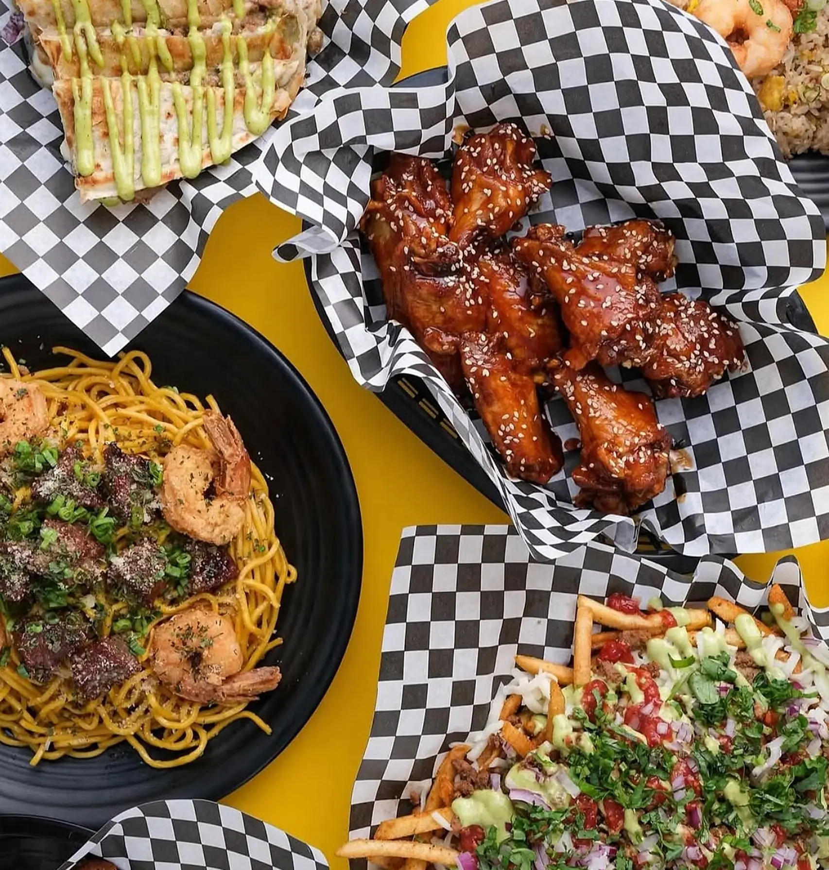 Overhead view of Nghbrs Kitchen dishes on black-checkered paper: sesame wings, garlic noodles with shrimp and steak, loaded fries, a grilled quesadilla and shrimp fried rice