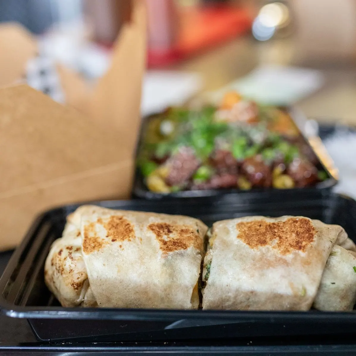 A grilled burrito cut in half in a black takeout tray, with a box of noodles behind it